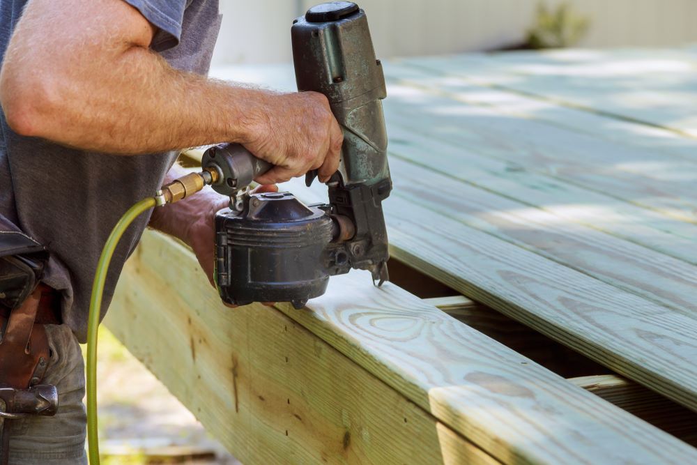 Worker using a nail gun to secure wooden boards during deck construction for a durable and safe structure.