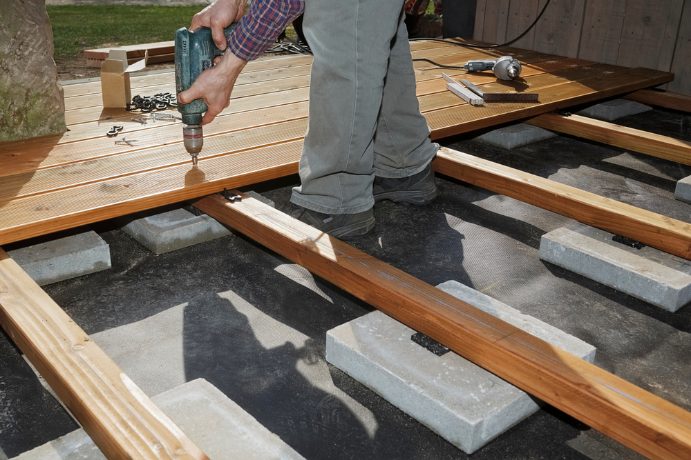 Close-up of a worker using a drill to secure wooden decking boards onto concrete foundation blocks during deck construction.