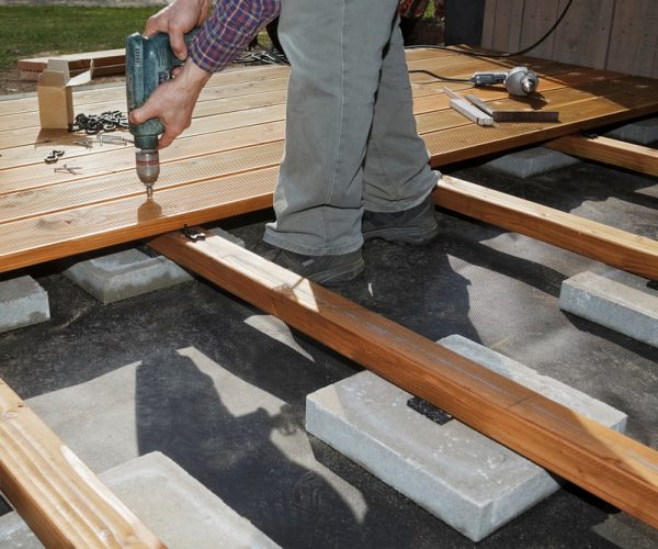 Close-up of a worker using a drill to secure wooden decking boards onto concrete foundation blocks during deck construction.
